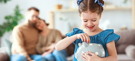 Young girl placing money in piggy bank with parents watching in the background.
